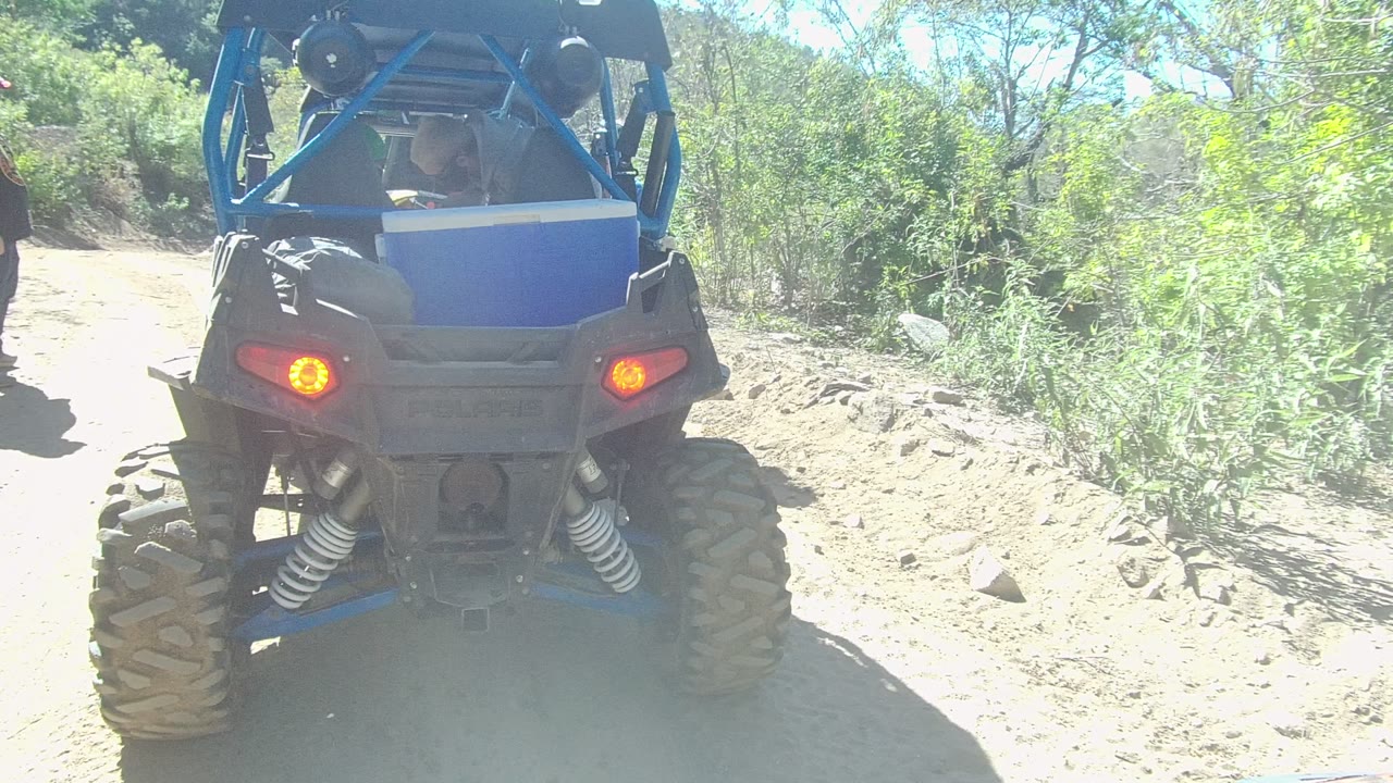 Rear view of a blue off-road vehicle on a dirt trail in Los Cabos, Baja California Sur, with red taillights, large tires, green brush, and a person partly entering at the left edge.