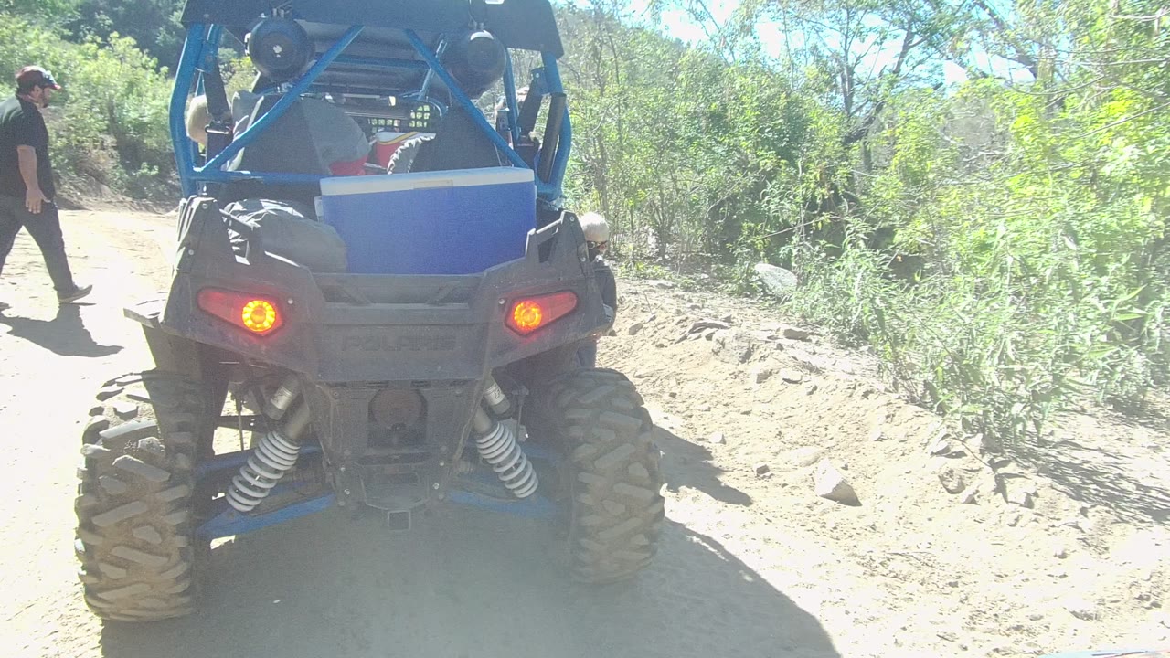 Person in dark clothing walking beside the rear of a blue off-road vehicle on a dusty path.