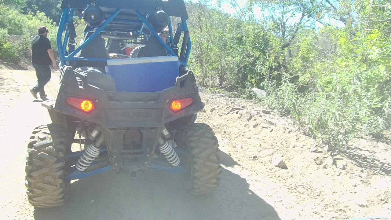 The person has moved farther right beside the blue off-road vehicle on the Los Cabos dirt path, while the vehicle rear, red taillights, brush, and rocks remain in view.