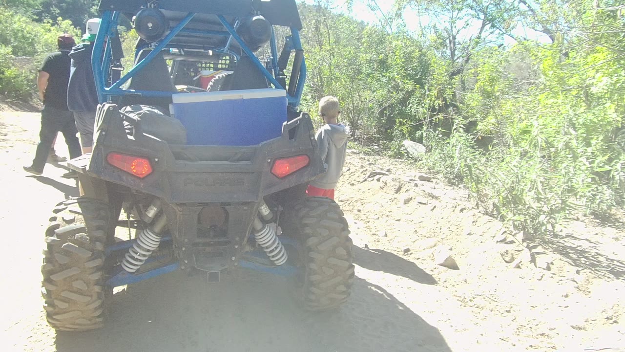 Rear view of a blue off-road vehicle stopped on a dirt trail in Los Cabos, Baja California Sur, with adults at left and a child near the passenger side.