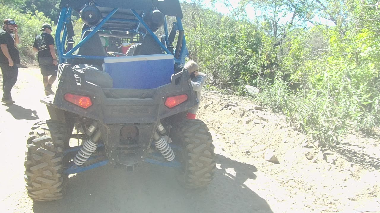 Rear composition of the blue off-road vehicle in Los Cabos with adults on the left side of the trail and a child partly visible near the right passenger area.