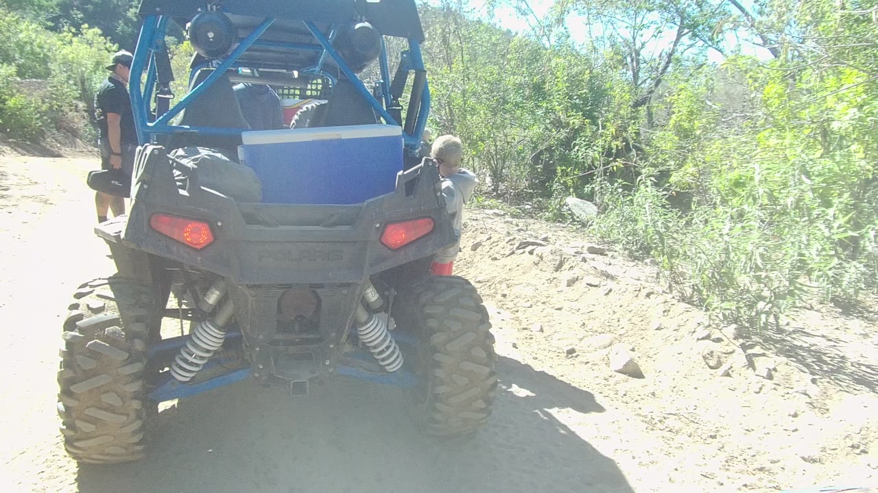 Rear view of a blue off-road vehicle parked on a rocky dirt path in Los Cabos, Baja California Sur, with an adult standing at left and a child-sized person near the right rear side.