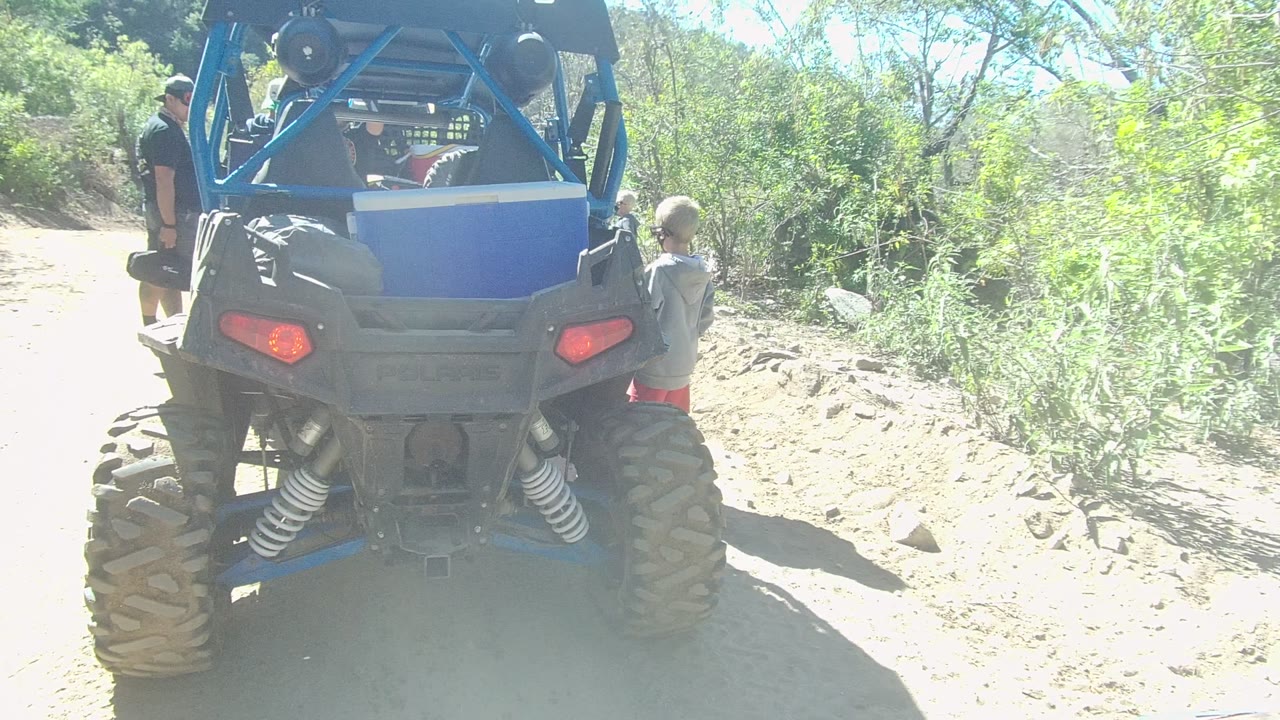 Blue off-road vehicle seen from behind on a bright dirt path with people at both sides.