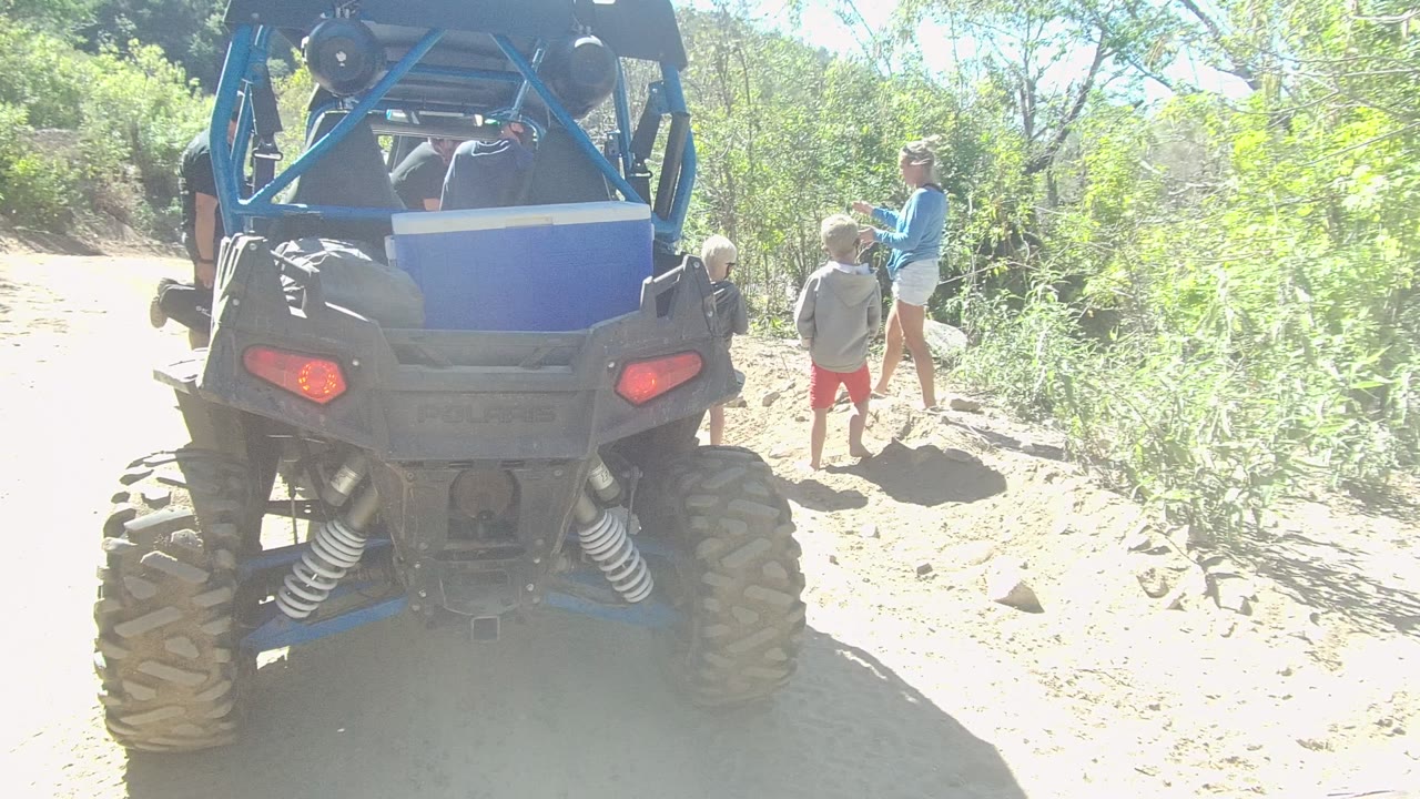 In Los Cabos, Baja California Sur, a blue off-road vehicle fills the left foreground while adults and children stand beside a dusty trail in bright sun.