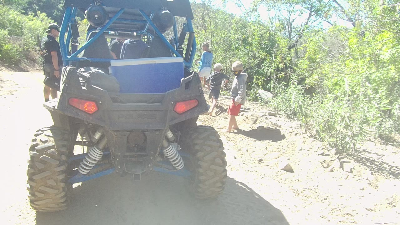 In Los Cabos, several people are clustered to the right of the blue off-road vehicle, including children near rocks and brush along the sunlit path.