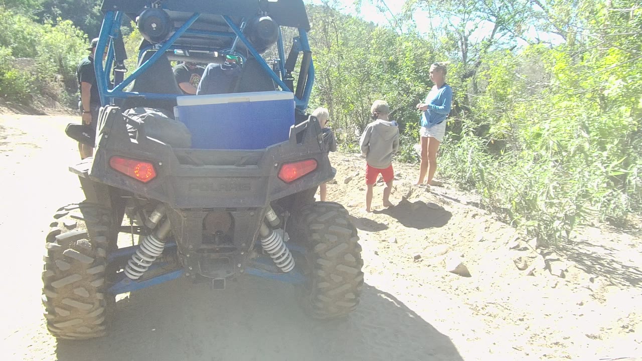 At the start of the Los Cabos clip, the rear of a blue UTV dominates the frame while a child in red shorts and an adult in a blue shirt stand to the right beside dry brush.