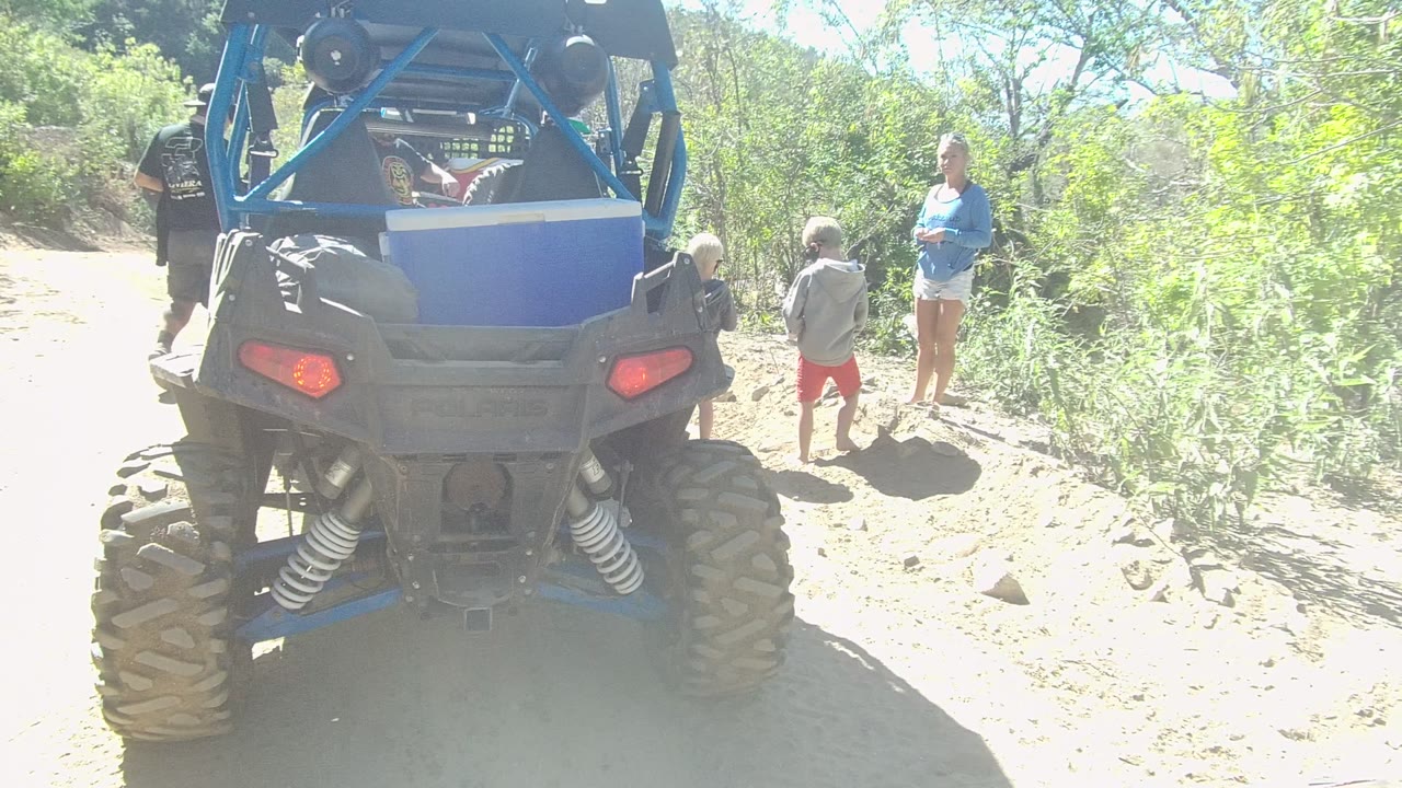 Rear view of a blue off-road vehicle on a Los Cabos dirt trail, with two children and an adult standing near bright roadside brush.