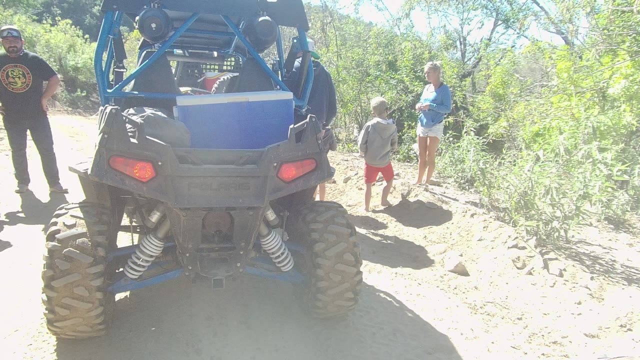 In Los Cabos, Baja California Sur, a blue off-road vehicle is parked on a dusty trail while a man in black, a child in red shorts, and a woman in blue stand near green brush.