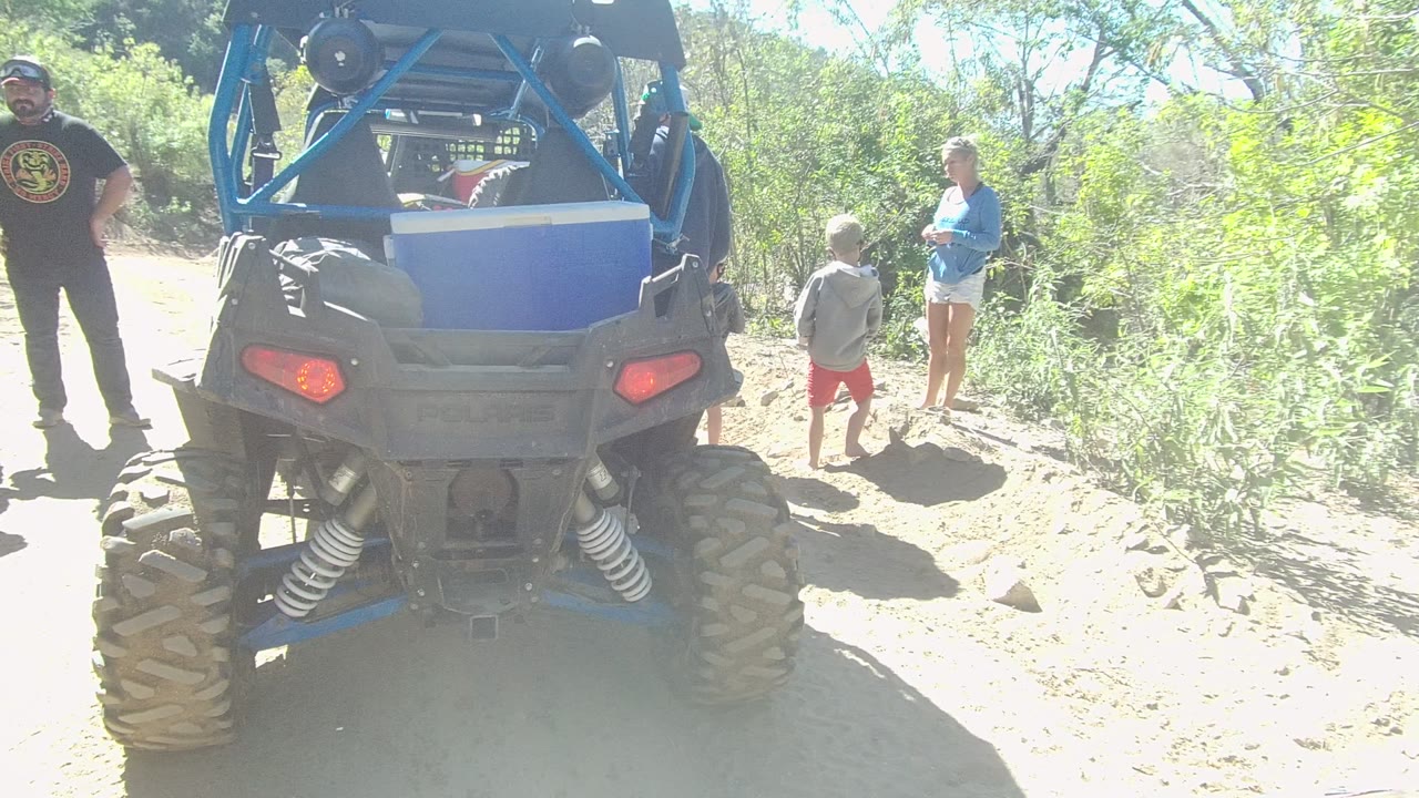 At 0:01 in Los Cabos, the rear of the blue off-road vehicle remains centered as the man in black stands to the left and the child and woman remain near the brush on the right.