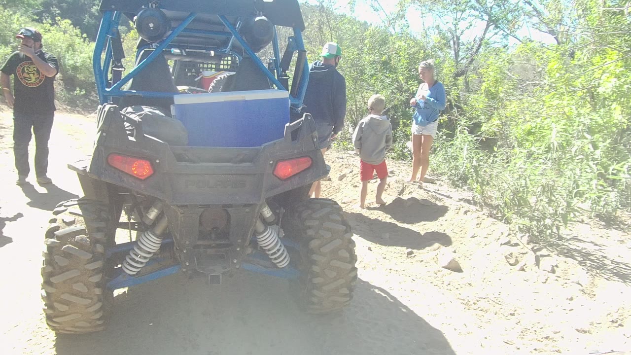 At 0:00 in Los Cabos, a blue off-road vehicle is seen from behind while adults and a child stand around it on a bright sandy trail lined with brush.