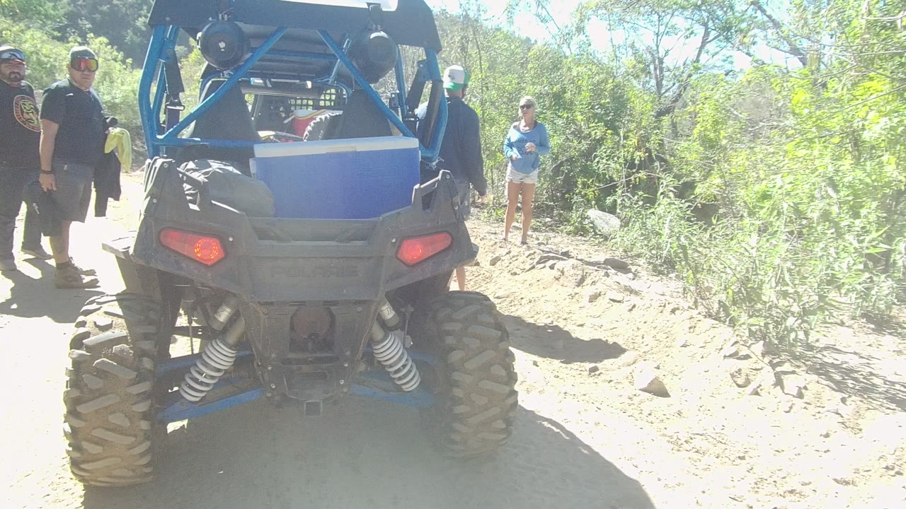 Rear view of a blue off-road vehicle on a dirt path in Los Cabos, Baja California Sur, with adults standing nearby in bright sun.