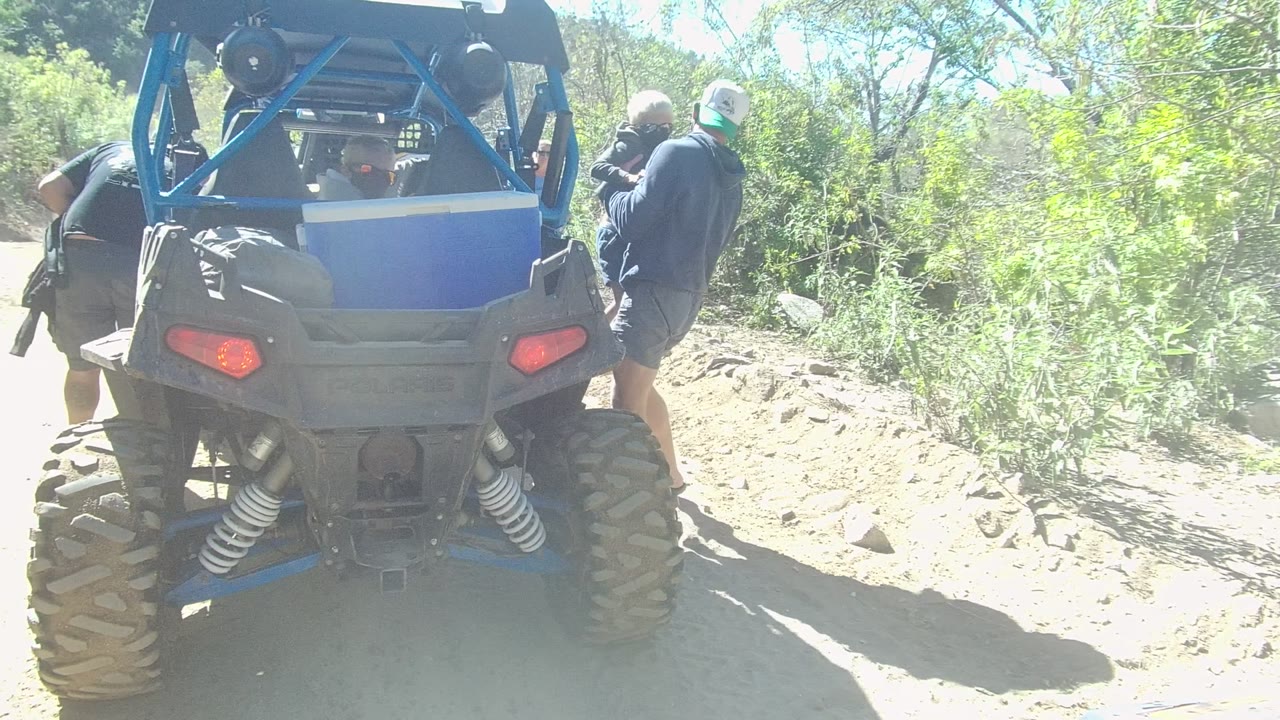 Rear view of a blue ATV in Los Cabos, Baja California Sur, with people partly visible beside it on a bright rocky dirt trail bordered by green brush.