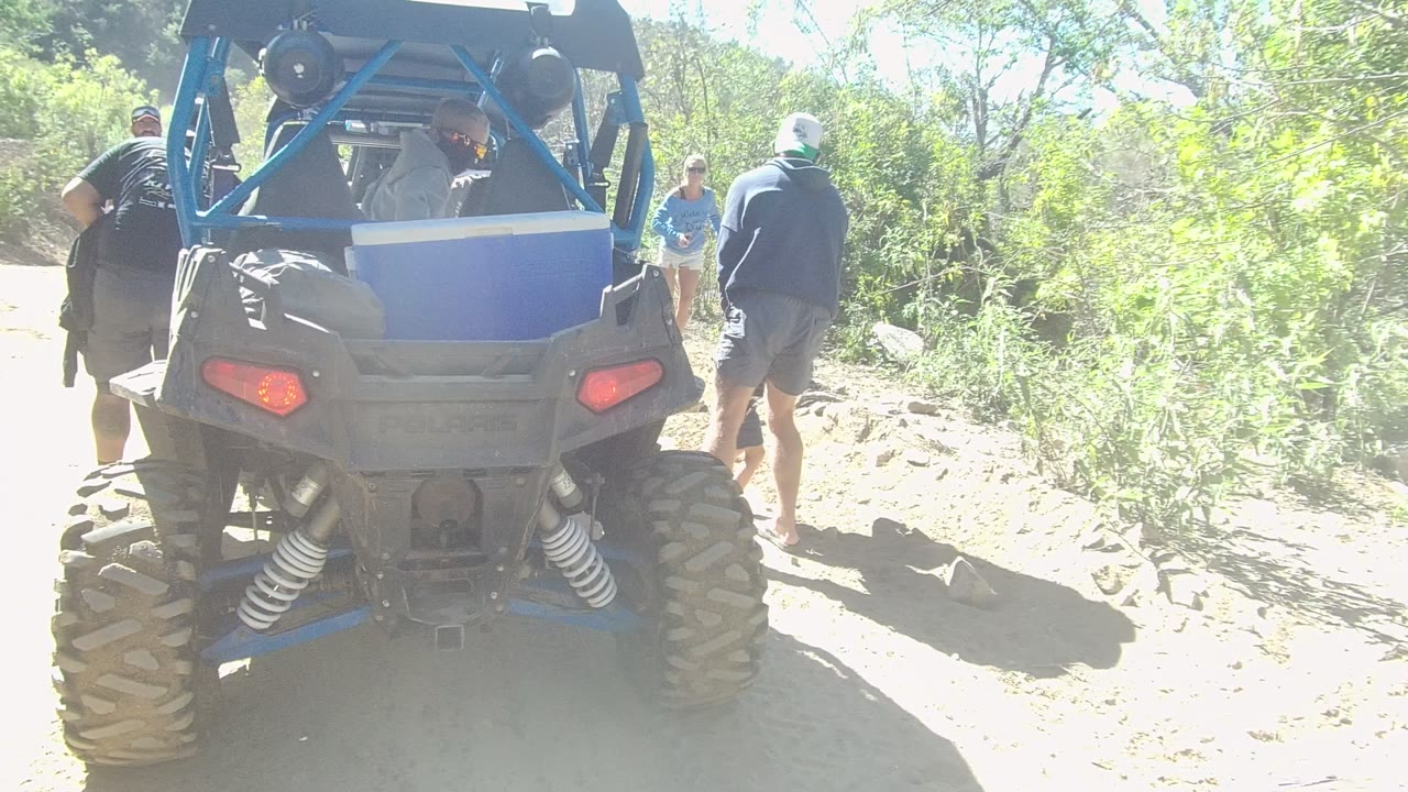 People walk around a blue ATV on a rocky trail in bright sun.
