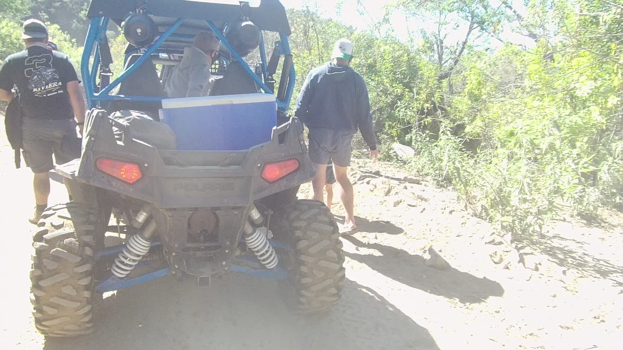 The ATV remains centered from behind in Los Cabos while two people move near the right side of the rocky path and bright green brush fills the background.