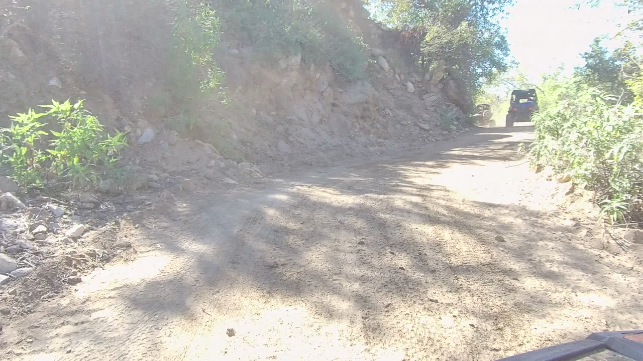 A narrow dirt road in Los Cabos, Baja California Sur curves between a rocky bank and green roadside vegetation, with a dark off-road vehicle small in the distance and no visible people.