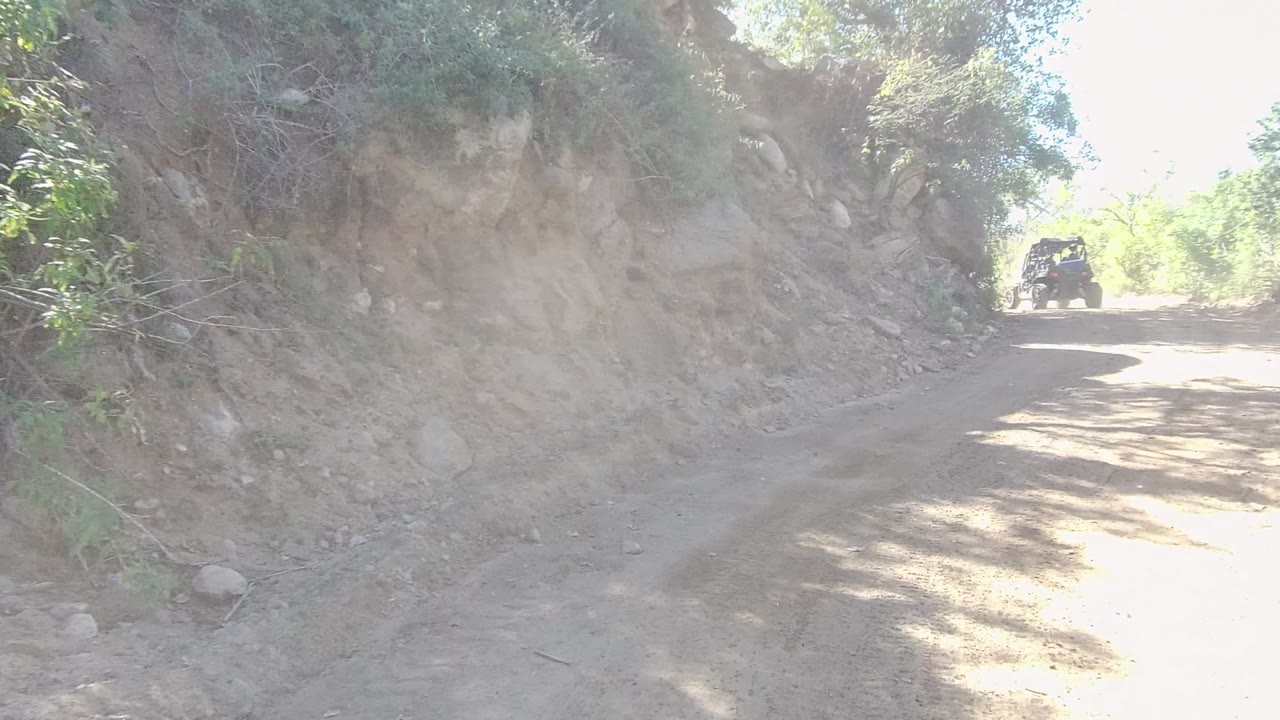 A dark off-road vehicle appears near the upper right of the Los Cabos trail while the dirt road curves around a steep rocky cut, with shrubs and no visible people.