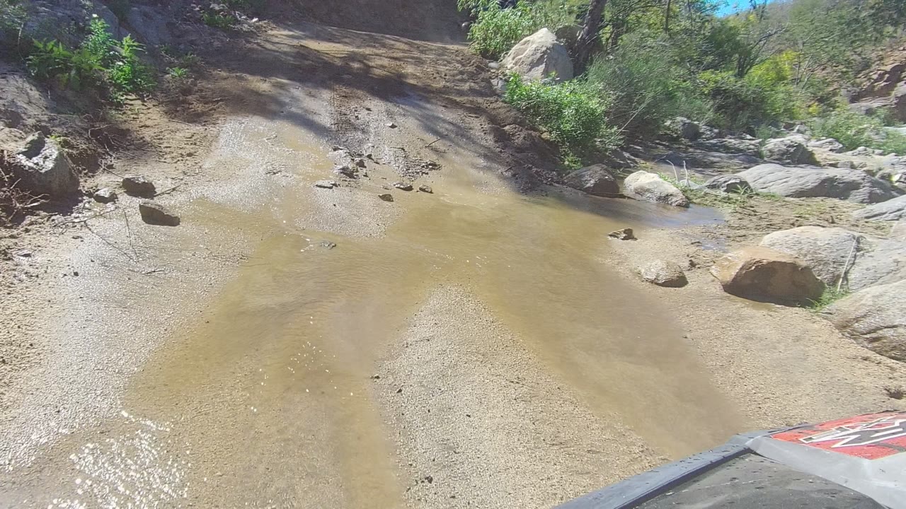 A forward vehicle view in Los Cabos looks across muddy water, pale gravel, boulders, brush, and a visible hood corner, with no visible people.