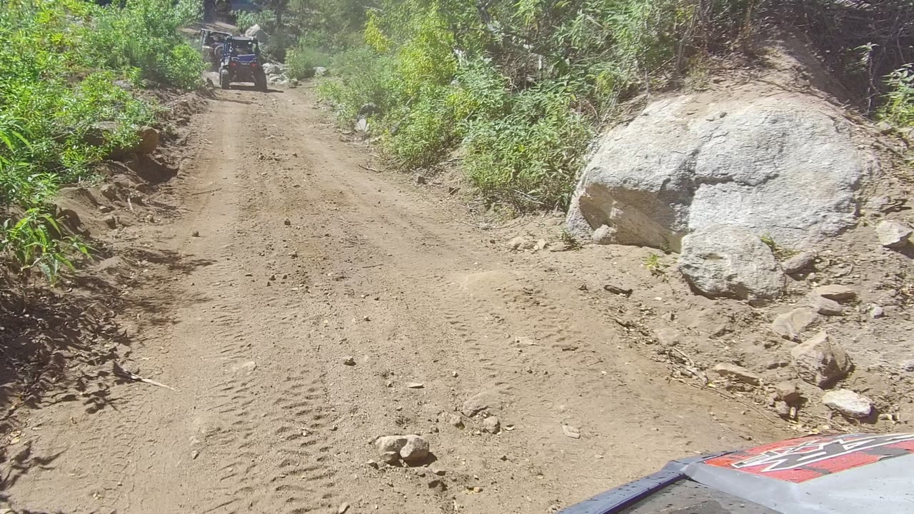 Forward view on a rocky dirt road in Los Cabos, Baja California Sur, with dense green brush on both sides, a large boulder to the right, an off-road vehicle ahead, and no visible people.