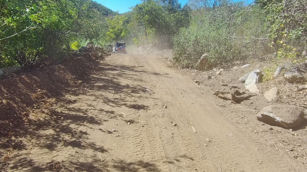 A distant off-road vehicle remains centered on a sunlit Los Cabos dirt road, with a thicker dust cloud, large rocks at the road edge, and no visible people.