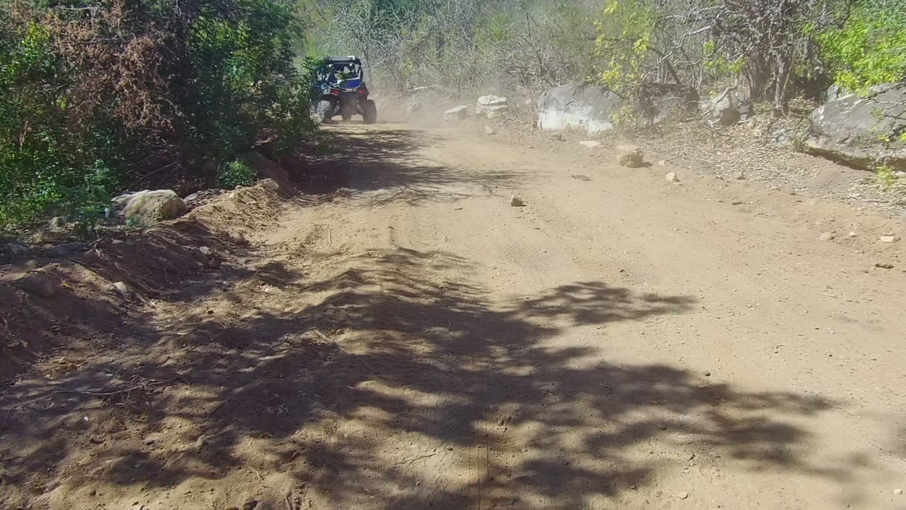 Dusty dirt road curves through brush with an off-road vehicle in the distance.