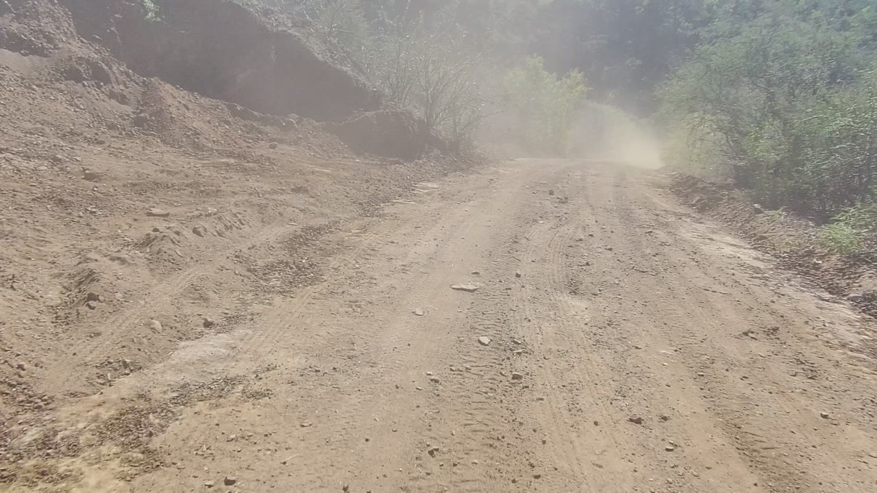 A tan dirt road in Los Cabos, Baja California Sur runs between rocky banks and sparse shrubs, with dust hanging ahead and no visible people.