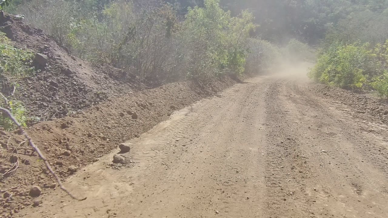 A rocky stretch of dirt road in Los Cabos passes a raised mound of earth and green brush, with dust visible down the road and no visible people.