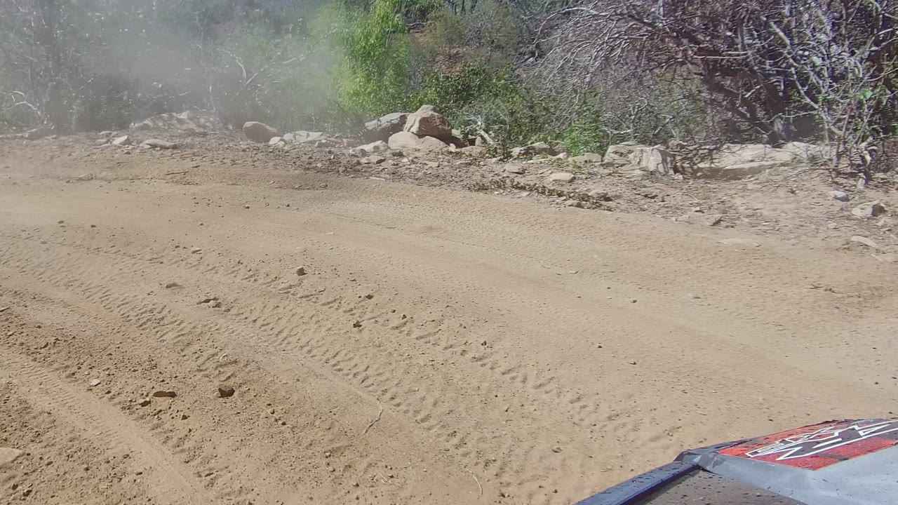 At 0:00 in Los Cabos, Baja California Sur, a sandy dirt road curves through rocky scrub terrain with a partial vehicle hood at the lower edge and no visible people.