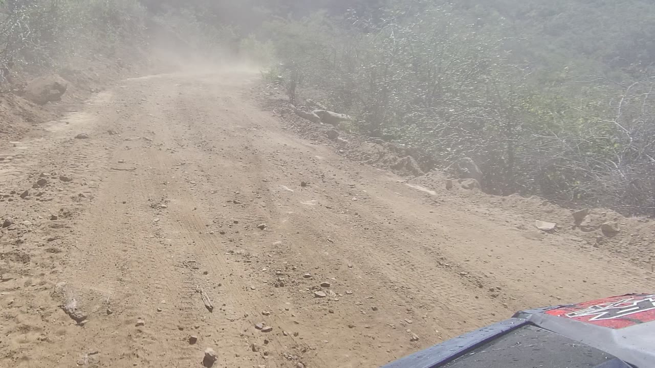 At 0:02 in Los Cabos, Baja California Sur, a rutted dirt road rises through dry brush and rocky shoulders while dust softens the background and no visible people appear.