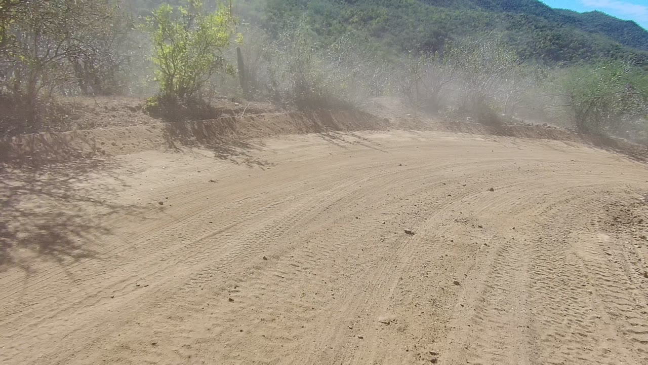 Wide view of a dusty sandy road in Los Cabos, Baja California Sur, with tire marks, roadside scrub, distant hills, bright sun, and no visible people.