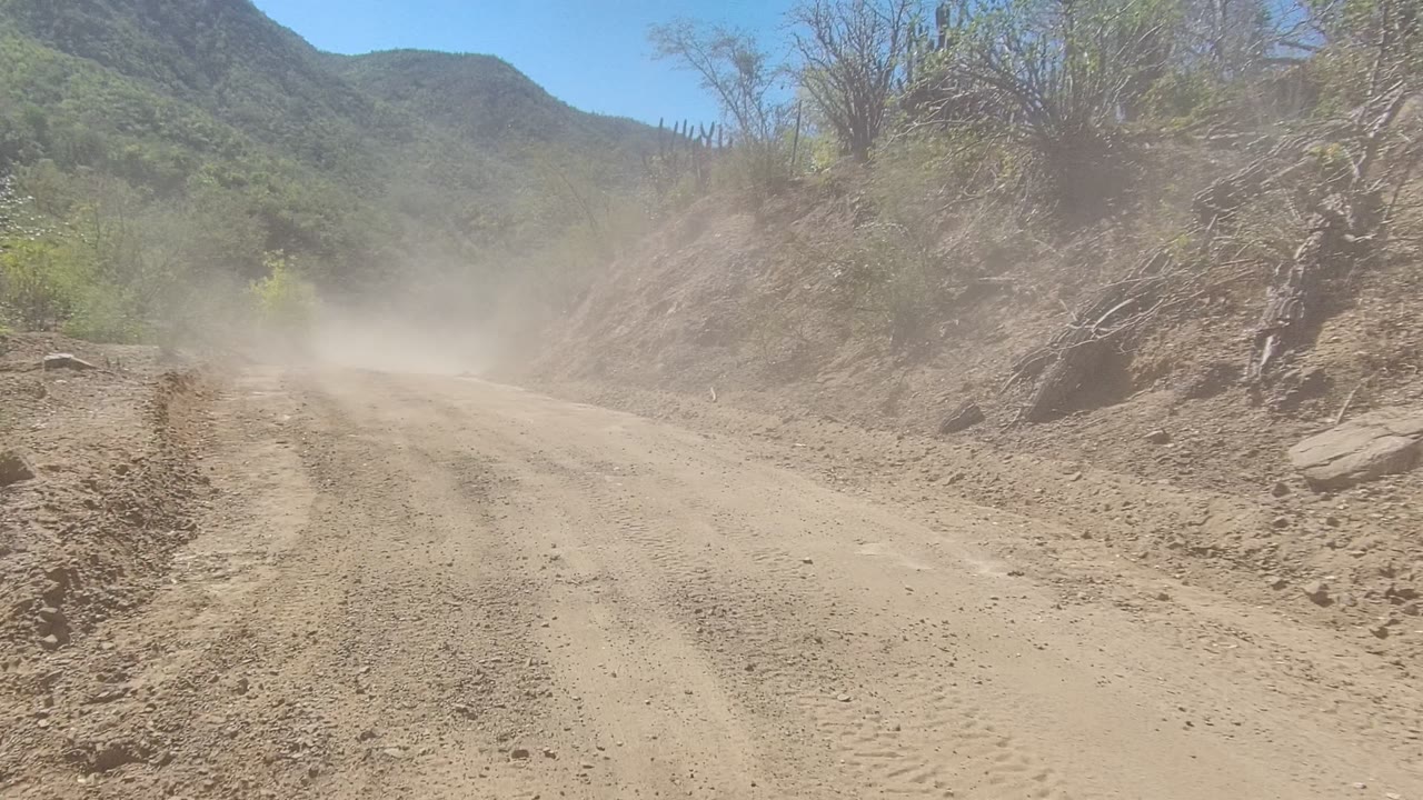 Dust rises over a dirt mountain road in Los Cabos, Baja California Sur, with rocky banks, scrub vegetation, distant hills, and no visible people.