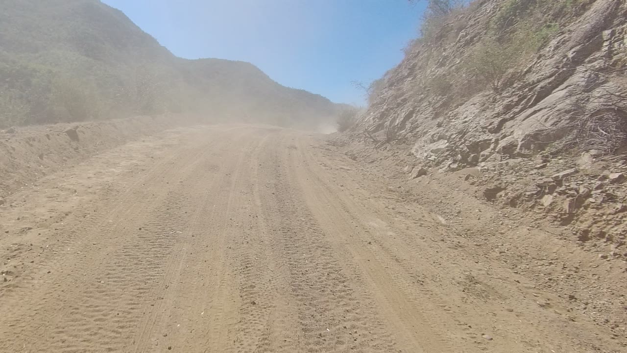 Los Cabos dirt road with tire tracks leading between rocky hills, a light dust haze ahead, blue sky above, and no visible people.
