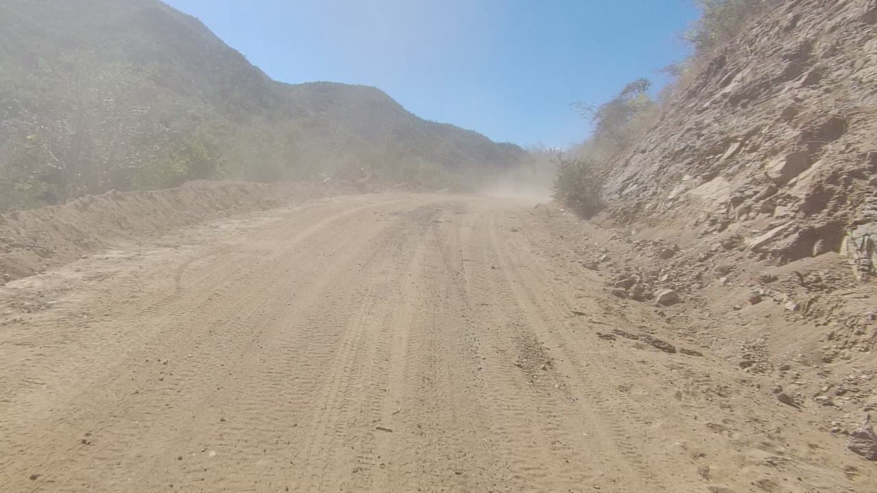 Unpaved road curving past a rocky cut bank with dust ahead