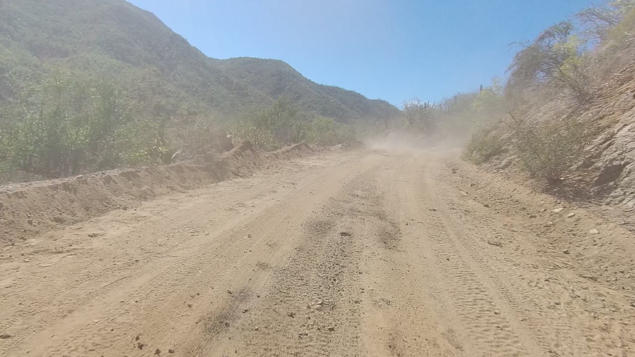 Los Cabos dirt track passing a rocky shoulder and green brush, with a dust plume softening the road ahead and no visible people.