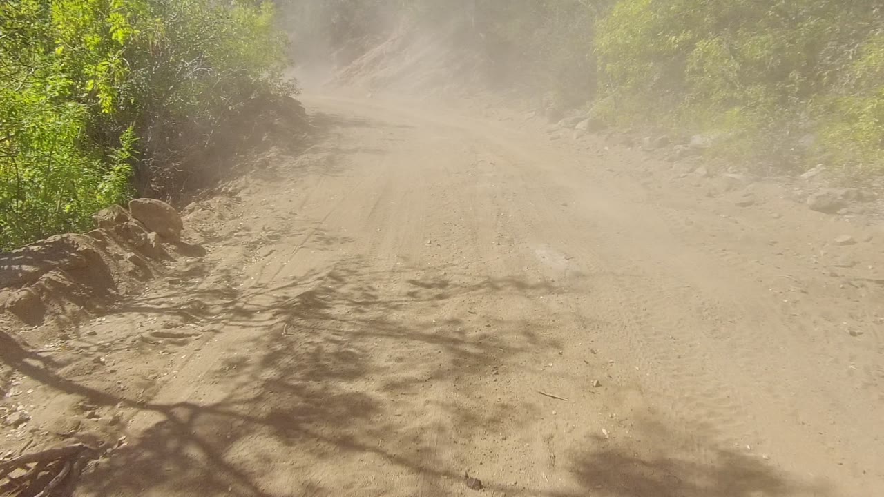 Opening view of a dusty dirt road in Los Cabos, Baja California Sur, with green brush on the left, rocky hillside on the right, airborne dust ahead, and no visible people.