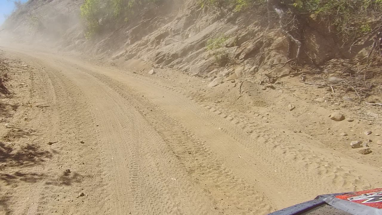 A partial vehicle hood enters the lower right as the Los Cabos dirt road bends past a rocky slope, with dust hanging over the track and no visible people.