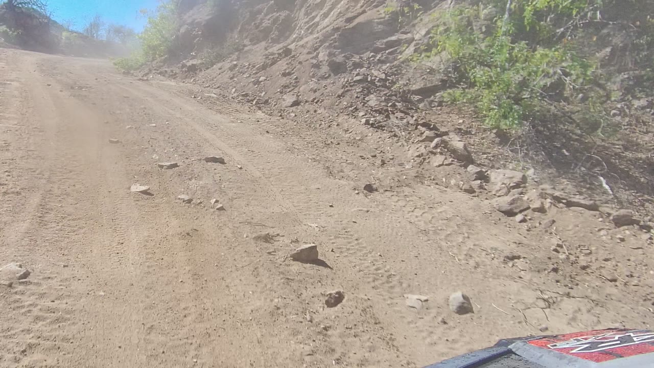 A forward road view in Los Cabos shows a dusty unpaved track with loose rocks, rocky banks, sparse shrubs, bright sky, and a partial vehicle hood at the lower right, with no visible people.