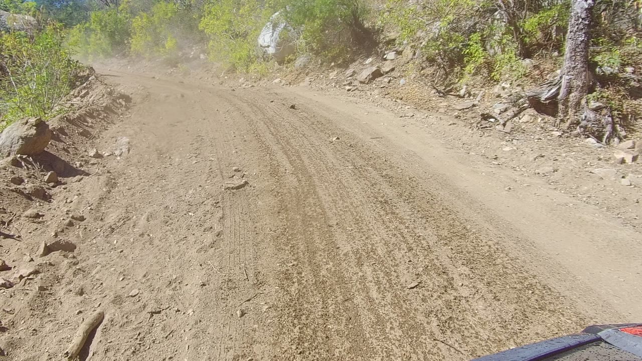 Rocky dirt road with dust ahead and brush along the shoulder.