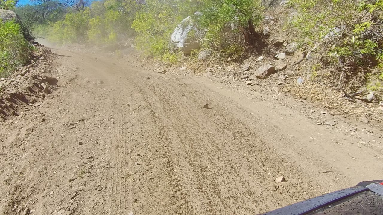 The road bends through rocky Los Cabos terrain with scrubby green vegetation, a bright blue sky patch, dust haze ahead, and no visible people.