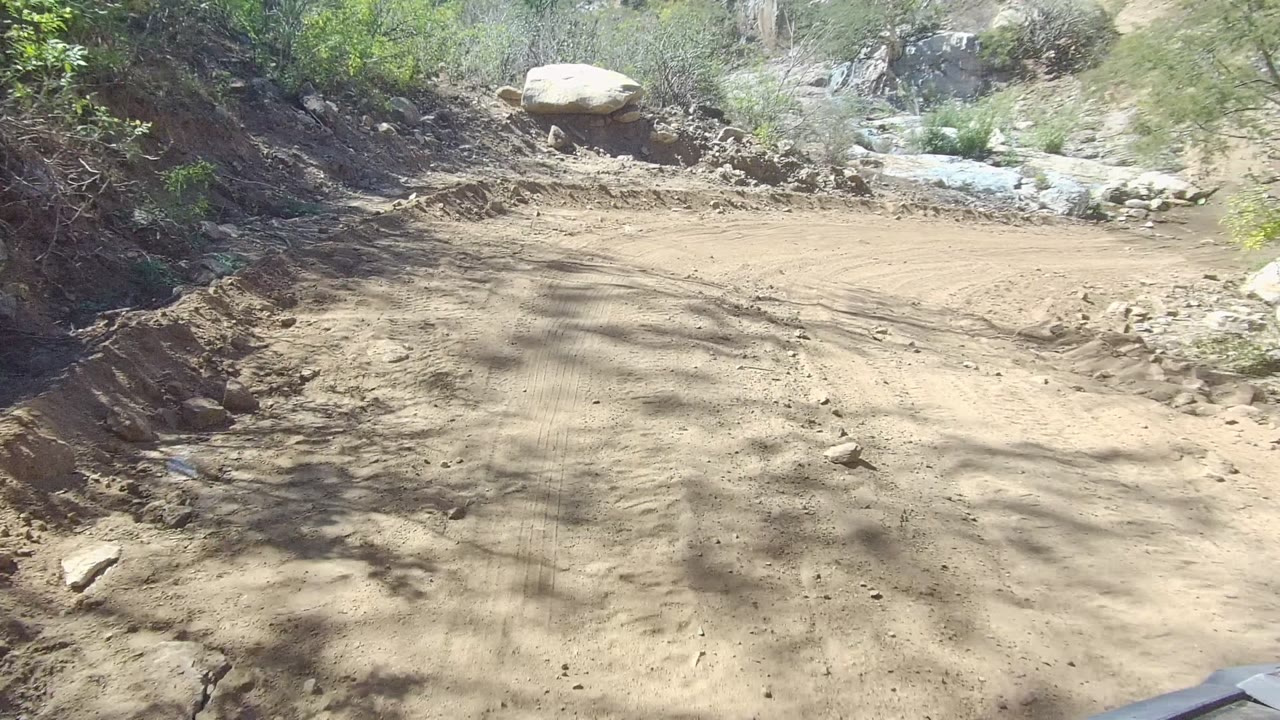 A sandy dirt road in Los Cabos, Baja California Sur runs past rocks and dry brush under bright light, with no visible people.