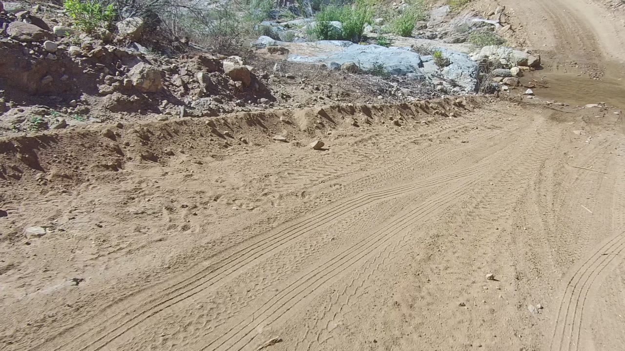 A rutted sandy road in Los Cabos passes a churned rocky berm and dry brush, with the water crossing still ahead and no visible people.