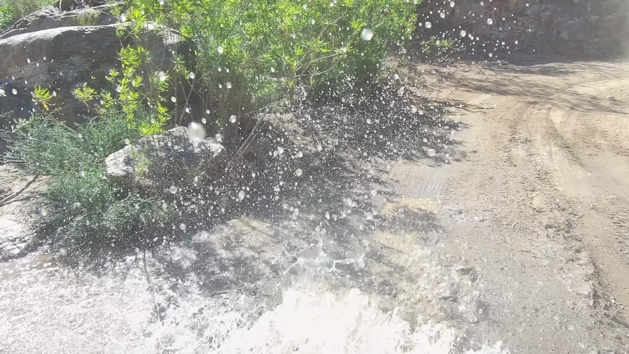 A large burst of water spray rises beside the dirt road in Los Cabos, Baja California Sur, with scrub vegetation, muddy water, rocks, and no visible people.