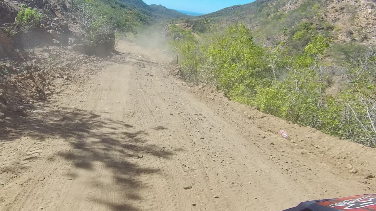 A rocky cut rises beside the unpaved Los Cabos road as the track continues between shrubs and distant hills, with no visible people.