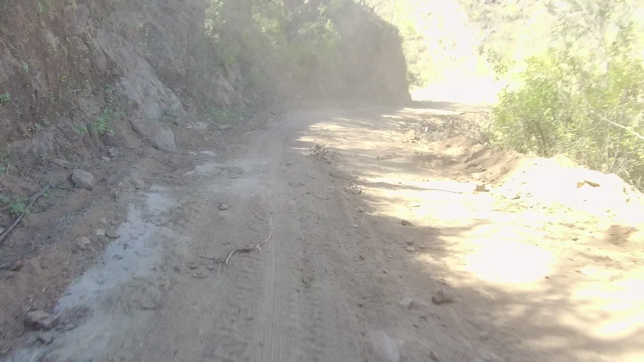 A narrow dirt road in Los Cabos, Baja California Sur runs between rough cut earth banks with bright sun ahead and no visible people.