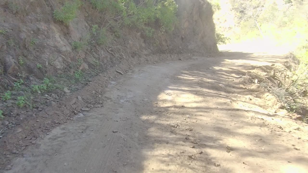 Dusty road curving beside a rocky slope with brush.