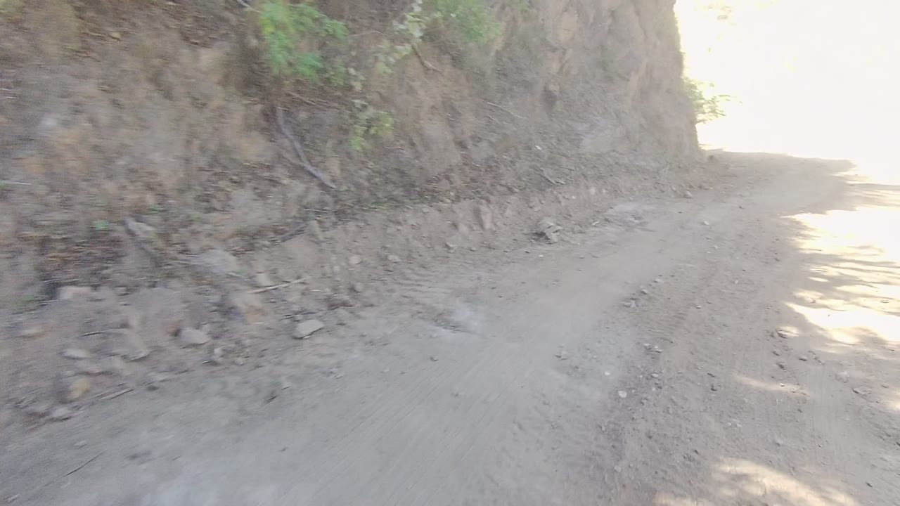 A close road-level view shows the unpaved Los Cabos route passing a steep dirt wall toward a bright sunlit bend, with no visible people.