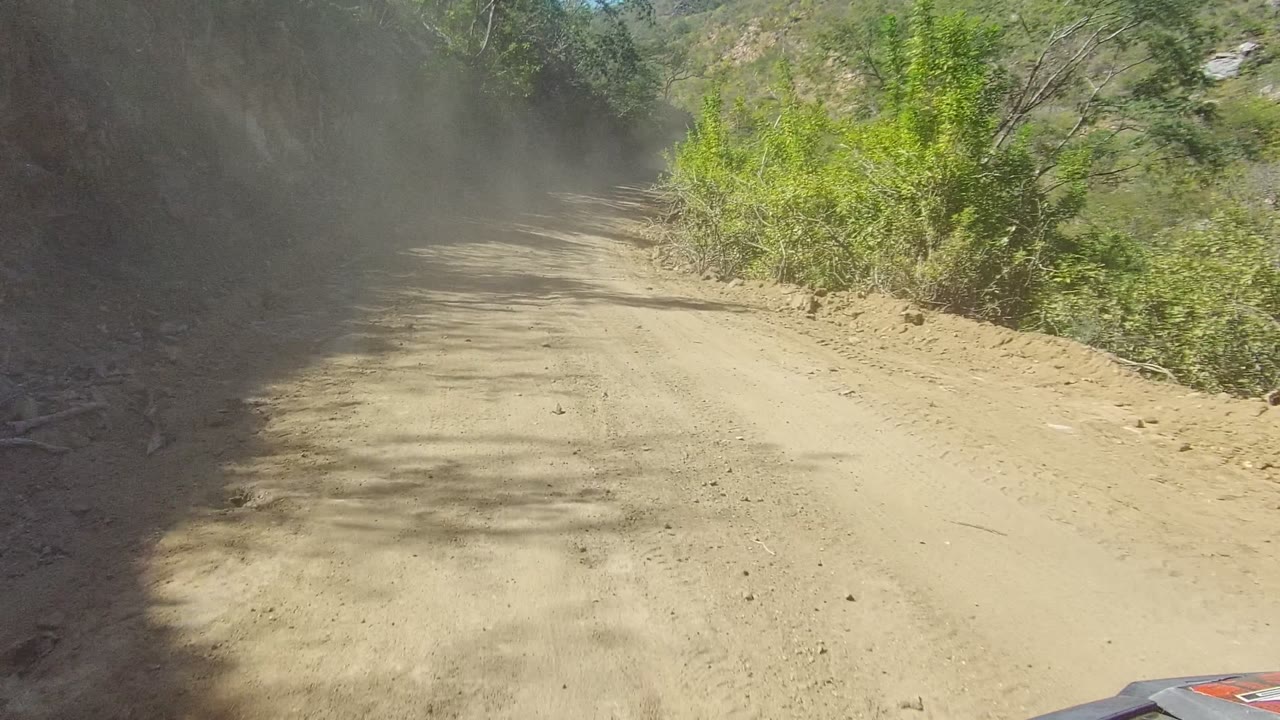 Vehicle-height view of a dusty dirt road in Los Cabos curving between a shaded rocky slope and green roadside brush, with no visible people.