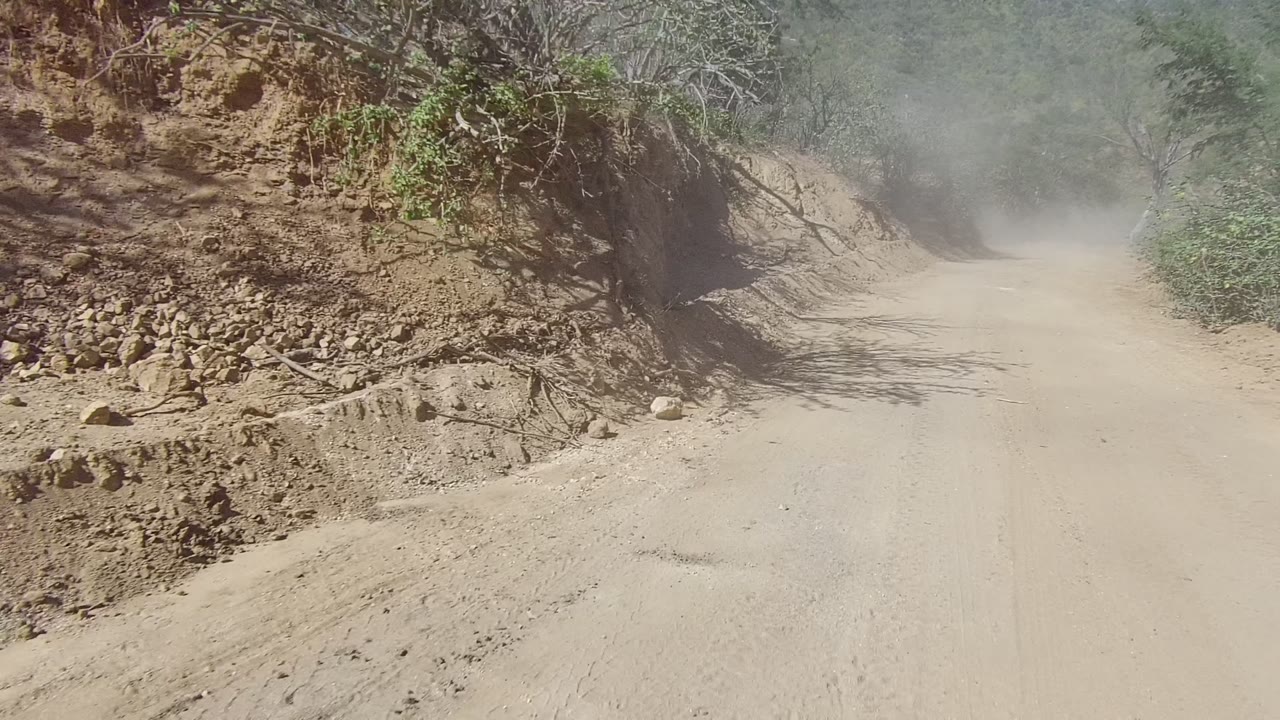 At 0:00, a dusty dirt road in Los Cabos, Baja California Sur curves past a rocky embankment with sparse brush and no visible people.