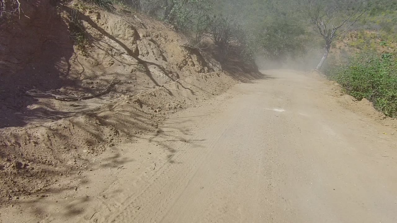 Pale dirt road running beside an eroded hillside with dust in the distance.