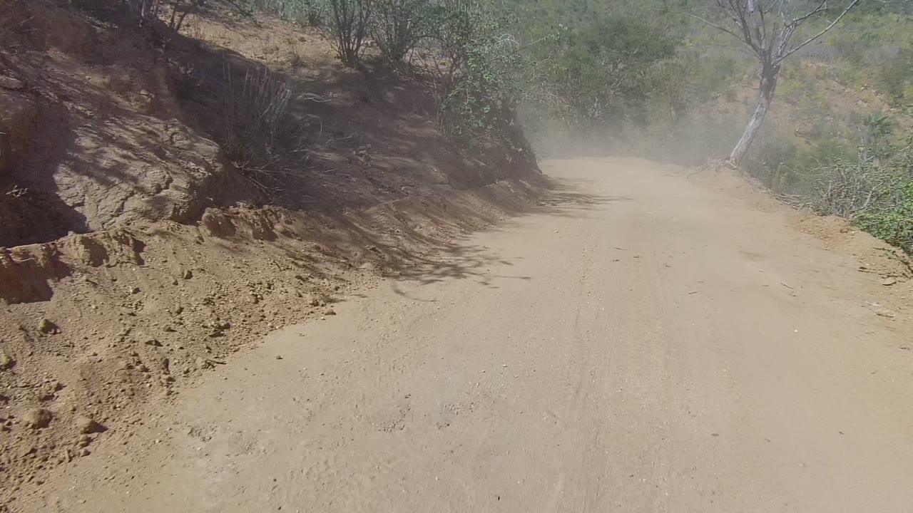 At 0:02, the road opens slightly in Los Cabos, with rocky banks, green shrubs, a dusty surface, dry hills beyond, and no visible people.
