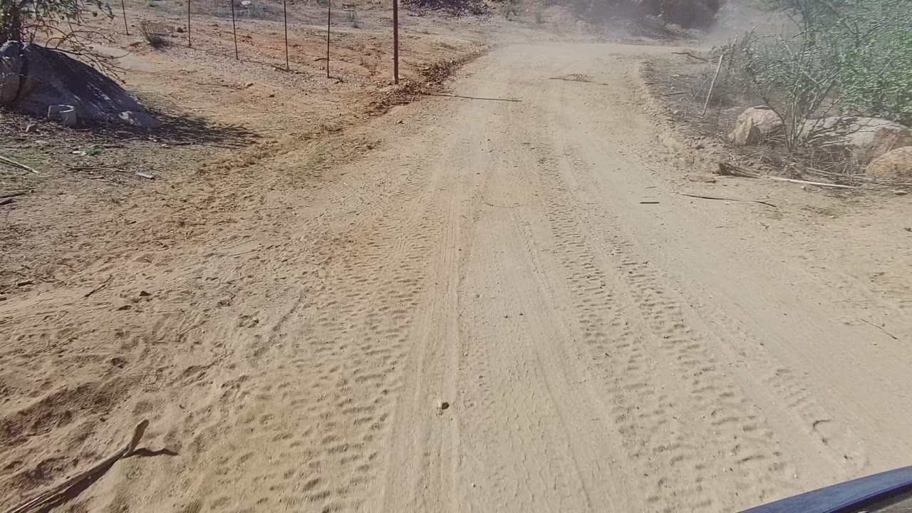 A closer forward view shows rutted sand, tire marks, dry roadside vegetation, a fence line, and no visible people in Los Cabos, Baja California Sur.